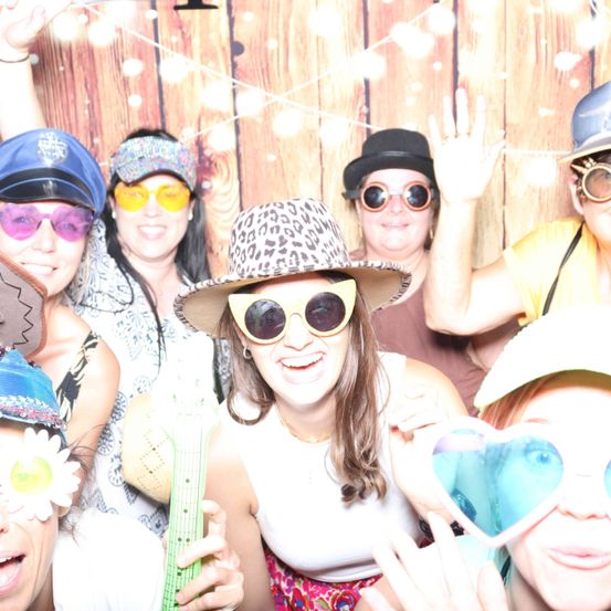 Eine Gruppe von Menschen in Hüten und Sonnenbrillen posiert für ein Foto in einer Fotokabine. Die Frau in der Mitte lächelt fröhlich.