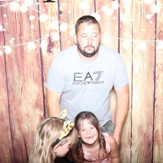 Ein Mann in einem Emporio Armani-Shirt steht hinter zwei kleinen Mädchen, von denen eines einen Leoparden-Kopfschmuck trägt, alle lächeln vor einem hölzernen Hintergrund.