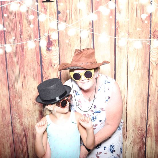 Eine Frau und ein junges Mädchen posieren vor einem hölzernen Hintergrund mit einer Lichterkette. Sie tragen beide Sonnenbrillen und Hüte. Die Frau trägt ein Blumenkleid und eine Halskette.