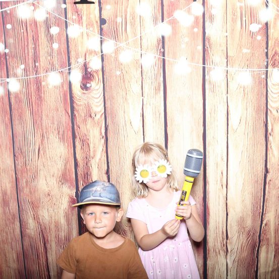 Zwei Kinder stehen vor einem hölzernen Hintergrund. Der Junge trägt einen Hut und das Mädchen eine Brille mit blumenförmigen Gläsern. Sie halten beide ein Mikrofon. Dahinter befindet sich eine Lichterkette.