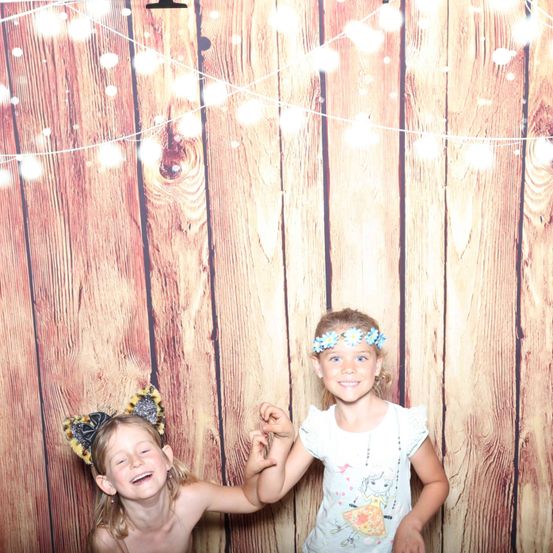 Zwei junge Mädchen stehen vor einem mit Lichterketten geschmückten Holzhintergrund. Das Mädchen links trägt Katzenohren und lächelt. Das Mädchen rechts trägt eine Blumenhaarschleife und hält die Hand des linken Mädchens.