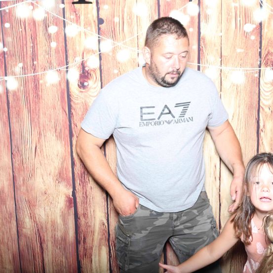 Ein Mann in einem grauen Emporio Armani-Shirt und Camouflage-Hosen steht hinter einer hölzernen Kulisse mit Lichterketten und posiert mit einem kleinen Mädchen in einem rosa Kleid.
