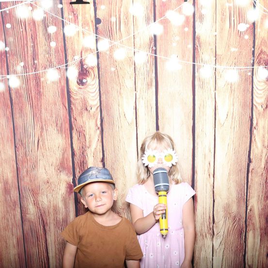 Zwei Kinder stehen vor einer Holzwand und lächeln für ein Foto. Der Junge trägt einen Hut und das Mädchen hält ein Mikrofon mit Sonnenblumen auf dem Kopf.