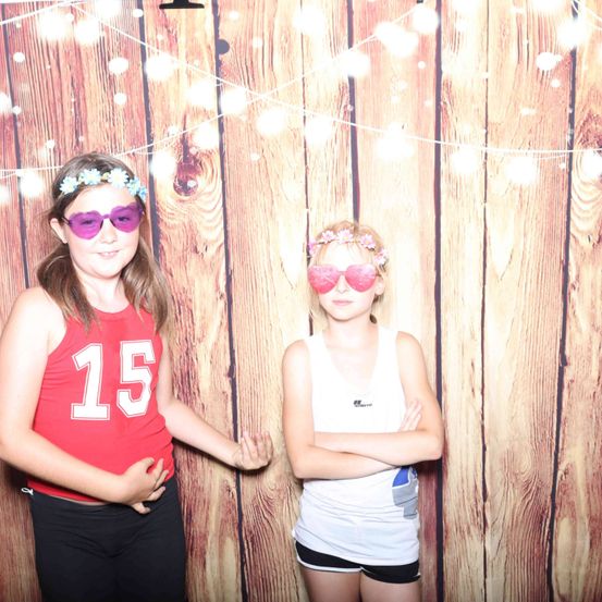 Zwei junge Mädchen mit lila Sonnenbrillen und Blumenkränzchen stehen nebeneinander vor einer Holzwand.