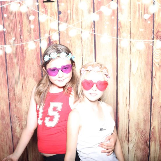 Zwei Mädchen stehen gemeinsam vor einer Holzwand, tragen herzförmige Sonnenbrillen und Blumenkränze und lächeln für ein Foto.