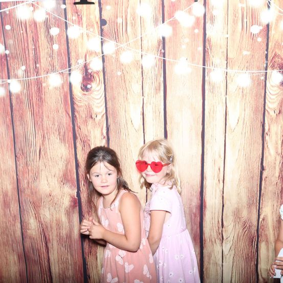 Zwei junge Mädchen posieren für ein Foto vor einer Holzwand mit Lichterketten. Beide tragen rosa Kleider.