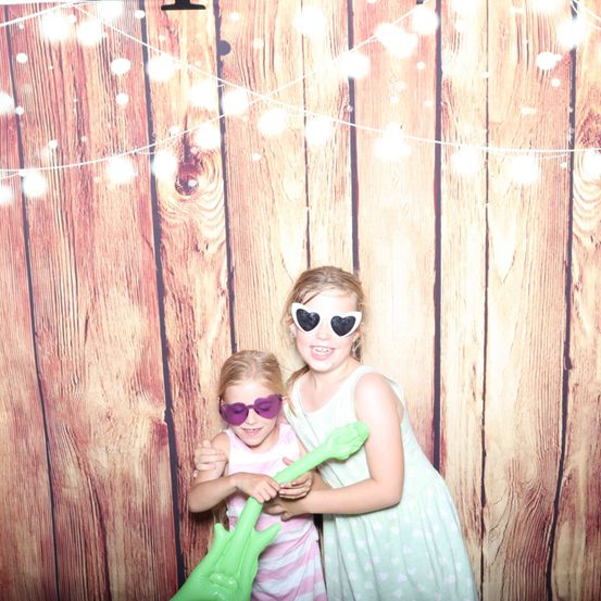 Zwei junge Mädchen posieren vor einem hölzernen Hintergrund mit Lichtern und tragen Sonnenbrillen. Eine hält eine grüne Gitarre, und beide sehen fröhlich aus.