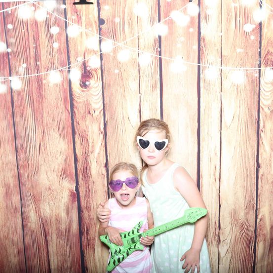 Zwei Mädchen in Sonnenbrillen und mit einer Gitarre posieren vor einem hölzernen Hintergrund mit Lichterketten.