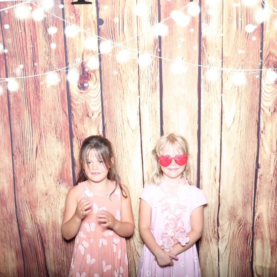 Zwei kleine Mädchen stehen nebeneinander vor einer mit Lichtern verzierten Holzwand. Das Mädchen auf der linken Seite trägt ein rosa Kleid mit Schmetterlingsdruck. Das Mädchen auf der rechten Seite trägt ein rosa Kleid mit Blumenkette und rote Sonnenbrille in Herzform.