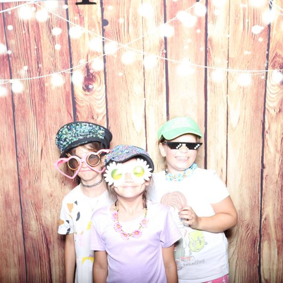 Drei Kinder in einem Fotostudio, jedes mit einzigartigen Accessoires: herzförmige Brille, eine blumenverzierte Mütze und eine grüne Kappe mit Sonnenbrille. Sie lächeln für die Kamera.