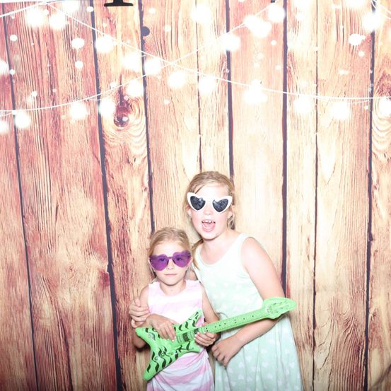 Zwei junge Mädchen, beide mit Sonnenbrillen, posieren mit einer grünen Gitarre vor einem hölzernen Hintergrund mit Lichterketten.