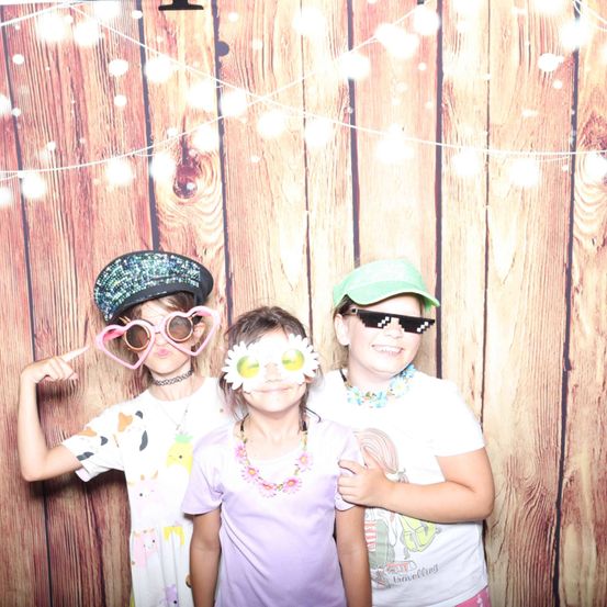 Drei Kinder posieren für ein Foto vor einem hölzernen Hintergrund und tragen lustige Brillen und Mützen. Das Mädchen in der Mitte trägt eine Blumenkopfbedeckung.