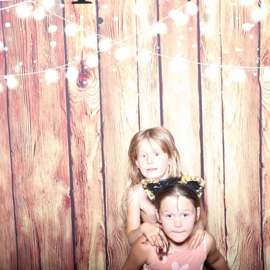 Zwei junge Mädchen posieren für ein Foto vor einem hölzernen Hintergrund mit Lichterketten. Das Mädchen auf der rechten Seite trägt Katzenohren.