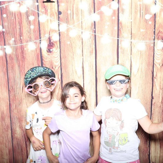 Drei Kinder stehen eng beieinander vor einem hölzernen Hintergrund mit Lichtern. Sie tragen Brillen und Hüte, und eines von ihnen hat eine Halskette.