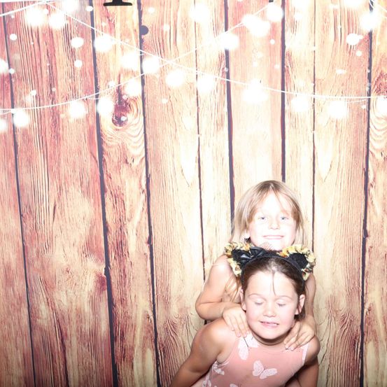Zwei junge Mädchen posieren für ein Foto vor einem hölzernen Hintergrund mit Lichterketten. Ein Mädchen steht auf den Schultern des anderen.