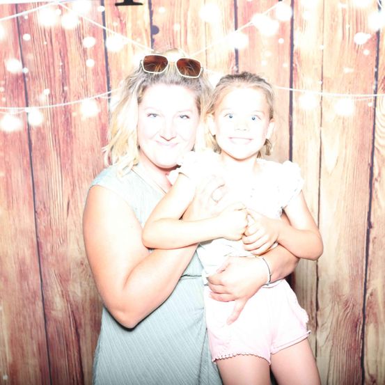 Eine lächelnde Frau hält ein kleines Mädchen in ihren Armen vor einem hölzernen Hintergrund. Beide lächeln.