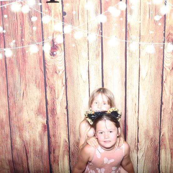 Zwei junge Mädchen posieren für ein Foto vor einem hölzernen Hintergrund mit Lichterketten. Ein Mädchen trägt ein Kleid mit Schmetterlingsdruck und eine Kopfband.