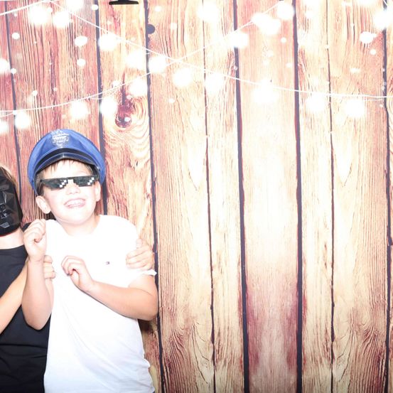 Ein junger Junge mit blauer Mütze und Sonnenbrille posiert vor einem hölzernen Hintergrund mit Lichterketten.