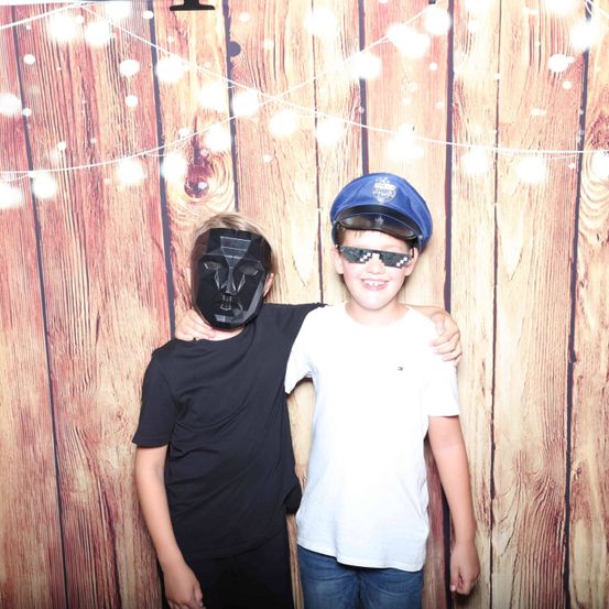 Zwei junge Jungen stehen eng beieinander in einem Fotoautomaten. Einer trägt eine schwarze Maske, der andere eine Polizeimütze und Sonnenbrille. Sie stehen vor einer Holzwand.