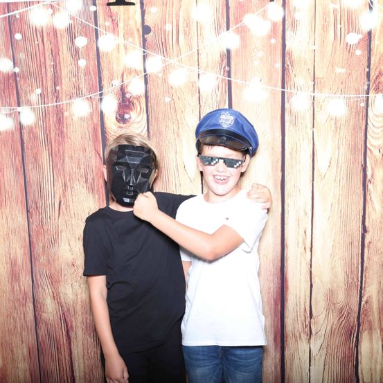 Zwei Jungen stehen vor einer Holzwand. Einer trägt eine schwarze Maske und schwarze Kleidung, während der andere ein weißes Shirt, Jeans und eine Polizeimütze trägt.