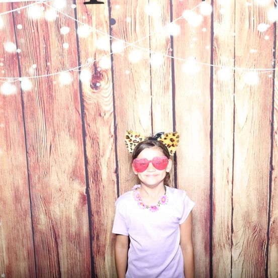 Ein kleines Mädchen steht vor einer Holzwand, trägt eine Leopardenkopfbedeckung, herzförmige Sonnenbrille und ein lila T-Shirt. Der Hintergrund zeigt eine festliche Lichterkette.