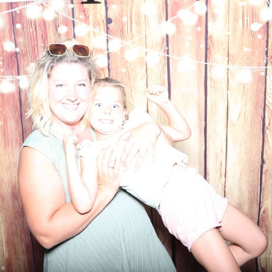 Eine Frau hält ein junges Mädchen in ihren Armen, beide lächeln. Sie stehen vor einem hölzernen Hintergrund mit Lichterketten.