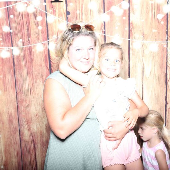Eine Frau mit Sonnenbrille hält ein kleines Mädchen mit rosa Shorts in den Armen. Ein weiteres kleines Mädchen steht hinter ihnen und alle lächeln vor einem hölzernen Hintergrund.