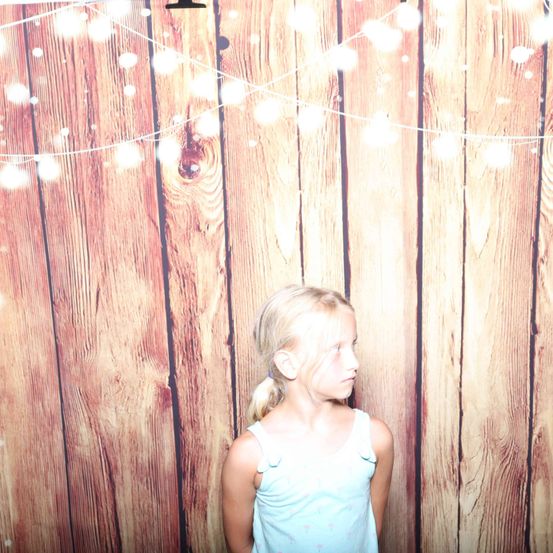 Ein kleines Mädchen mit einem Pferdeschwanz steht vor einer Holzwand mit Lichterketten. Sie schaut zur Seite, vielleicht für ein Foto.