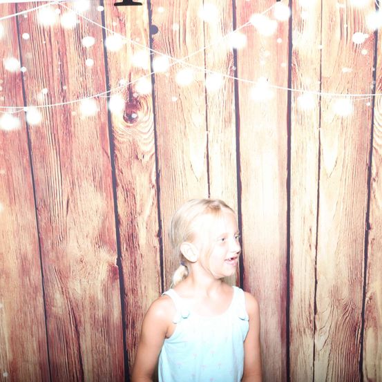 Ein kleines Mädchen mit blonden Haaren lächelt vor einem hölzernen Hintergrund mit dekorativen Lichtern.
