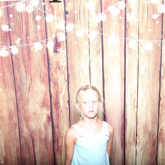 Ein junges Mädchen mit hellblondem Haar steht vor einem mit Lichterketten geschmückten Holzhintergrund. Sie trägt ein ärmelloses blaues Kleid.