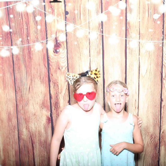 Zwei junge Mädchen stehen nebeneinander vor einem hölzernen Hintergrund mit Lichterketten. Ein Mädchen hat eine Leopardenkopfbedeckung, und beide haben ihre Zungen herausgestreckt.
