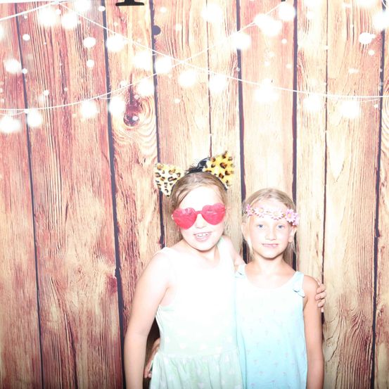 Zwei Mädchen stehen nebeneinander vor einem hölzernen Hintergrund mit Lichterketten. Eine trägt rote Gläser und ein Katzenohren-Kopfband. Die andere hat ein Blumen-Kopfband.