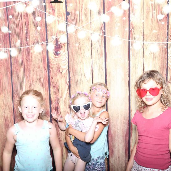 Vier junge Mädchen stehen vor einem hölzernen Hintergrund mit Lichterketten. Eines der Mädchen trägt eine herzförmige Sonnenbrille. Eine andere hält ein Baby und lächelt, während die anderen in die Kamera schauen.