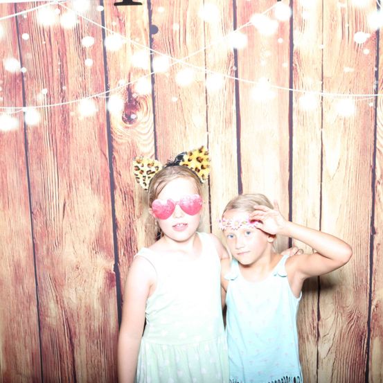 Zwei Mädchen stehen vor einem hölzernen Hintergrund, eines trägt herzförmige Brillen und einen Leopardenkopfschmuck, das andere einen Blumenkopfschmuck. Sie schauen in die Kamera.