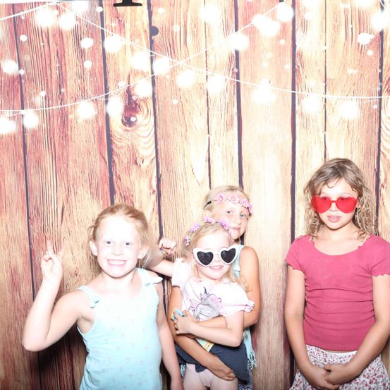 Vier junge Mädchen in Sommerkleidung stehen vor einem hölzernen Hintergrund. Die Mädchen lächeln und machen Friedenszeichen. Zwei der Mädchen tragen Sonnenbrillen.