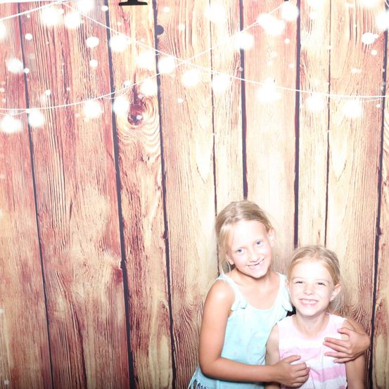 Zwei junge Mädchen posieren für ein Foto vor einem hölzernen Hintergrund mit Lichterketten. Sie lächeln und scheinen Spaß zu haben.