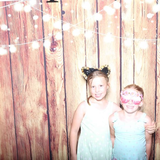 Zwei junge Mädchen stehen vor einer hölzernen Wand mit Lichtern. Sie tragen Kleider und lächeln. Das Mädchen auf der linken Seite hat Katzenohren-Kopfbänder und das Mädchen auf der rechten Seite hat rosa Brille.