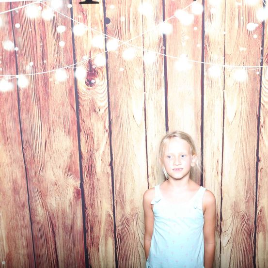 Ein junges Mädchen mit hellblondem Haar steht vor einer mit Lichterketten geschmückten Holzwand. Sie trägt ein hellblaues ärmelloses Oberteil.