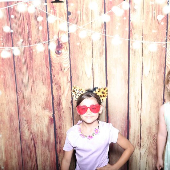 Ein kleines Mädchen mit roten Herzgläsern und einem Leoparden-Kopfband steht vor einem hölzernen Hintergrund mit Lichterketten. Sie lächelt, trägt ein lila Hemd und eine Halskette mit Blumendesigns.