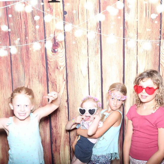 Vier junge Mädchen posieren für ein Foto vor einem hölzernen Hintergrund mit Lichterketten. Sie tragen bunte Accessoires und lächeln fröhlich.