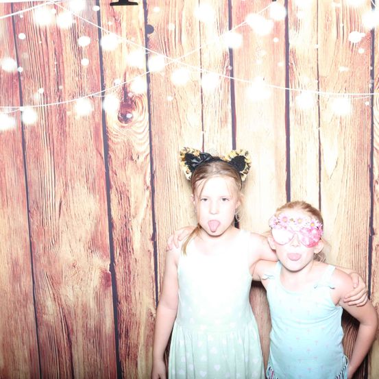 Zwei junge Mädchen posieren vor einem hölzernen Hintergrund, eine mit Katzenohren und die andere mit Blumen-Sonnenbrille.