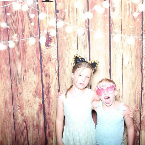 Zwei Mädchen posieren für ein Foto mit einem hölzernen Hintergrund und Lichterketten, eines trägt eine Katzen-Kopfbedeckung und das andere trägt blumenbedruckte Brille.