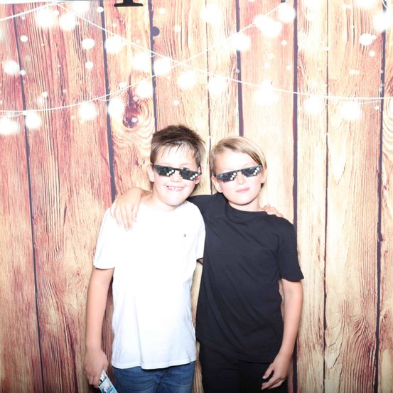 Zwei Jungen stehen vor einer Holzwand, beide tragen Sonnenbrillen. Der Junge links trägt ein weißes T-Shirt und Jeans, der Junge rechts ein schwarzes T-Shirt und schwarze Hose.