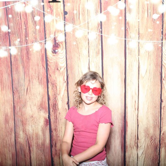Ein junges Mädchen mit herzförmigen Sonnenbrillen und einem rosa T-Shirt steht vor einem hölzernen Hintergrund mit dekorativen Lichtern.