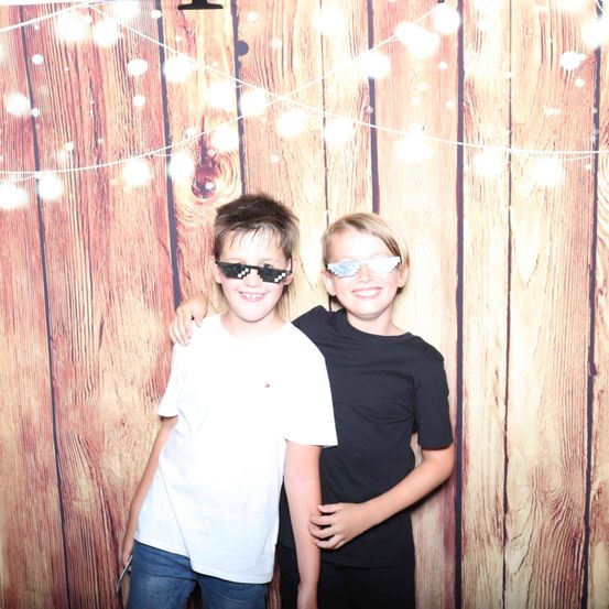 Zwei junge Jungen stehen vor einem hölzernen Hintergrund mit Lichtern. Einer trägt ein weißes T-Shirt und Jeans, der andere ein schwarzes T-Shirt. Beide tragen Sonnenbrillen.
