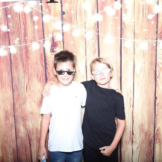 Zwei Jungen, einer in einem weißen T-Shirt und der andere in einem schwarzen T-Shirt, stehen eng beieinander vor einem hölzernen Hintergrund mit dekorativen Lichtern. Sie tragen beide eine Sonnenbrille.