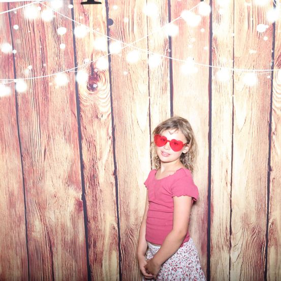 Ein kleines Mädchen mit roten Herz-förmigen Sonnenbrillen und einem rosa Oberteil steht vor einer mit Lichtern geschmückten Holzwand.