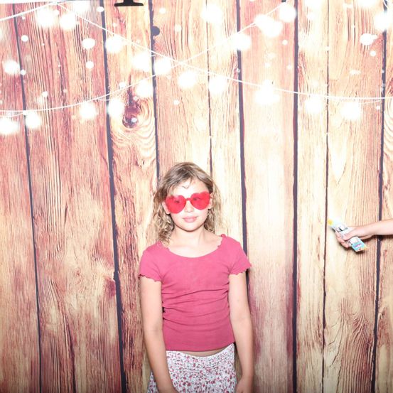 Ein junges Mädchen steht vor einer Holzwand mit Lichterketten. Sie trägt ein rosa T-Shirt und herzförmige Sonnenbrille. Hinter ihr hält eine Person eine Flasche Haarspray.
