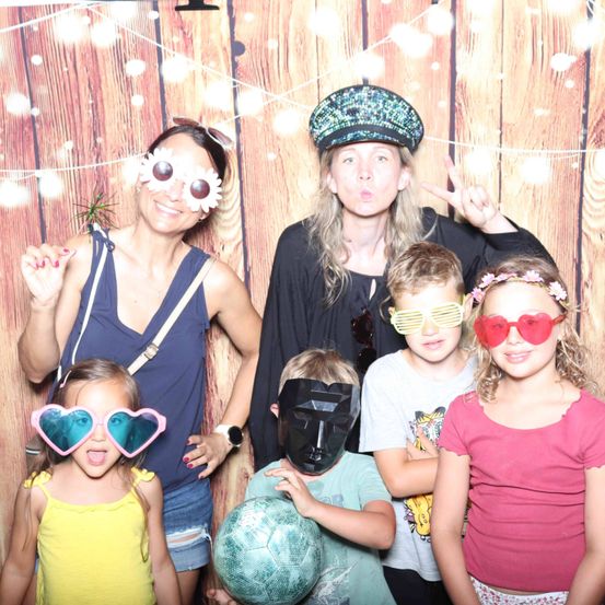 Eine Gruppe von Menschen mit Sonnenbrillen, die vor einer Holzwand für ein Foto posieren. Ein Mädchen in Gelb trägt herzförmige Sonnenbrillen.