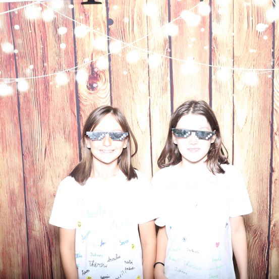 Zwei junge Mädchen stehen nebeneinander und tragen Sonnenbrillen mit pixeligen Gläsern. Sie lächeln und posieren für ein Foto vor einem hölzernen Hintergrund mit dekorativen Lichtern.